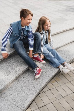 full length of cheerful well dressed children sitting on stairs of urban street 