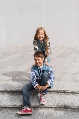 cheerful girl in stylish clothes leaning on shoulders of happy preteen boy sitting on stairs  clipart