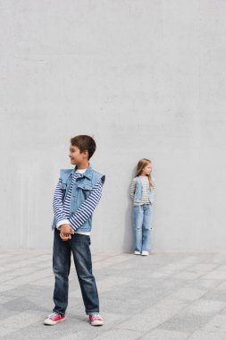 full length of cheerful boy in stylish denim outfit standing near girl on blurred background  clipart