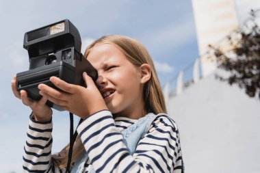 Düşük açılı kot yelekli ve çizgili uzun kollu tişörtlü bir kız klasik fotoğraf makinesinin fotoğrafını çekiyor. 