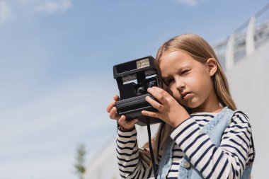 preteen girl in denim vest and striped long sleeve shirt taking photo of vintage camera 