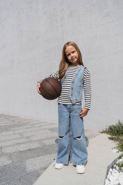 Kot yelekli ve kot pantolonlu bir kız basketbol topunu alışveriş merkezinin yakınında tutuyor. 