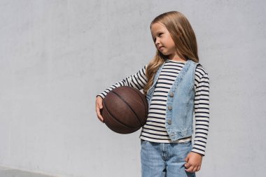 preteen girl in denim vest and blue jeans holding basketball near mall building 
