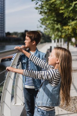 stylish kids in denim vests standing near metallic fence on riverside clipart