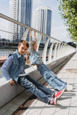 well dressed kids in denim vests and jeans posing near metallic fence on riverside clipart