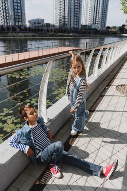 high angle view of well dressed kids in denim vests and jeans posing near metallic fence on embankment of river  clipart