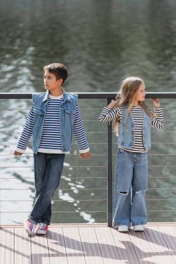 full length of well dressed kids in denim vests and jeans posing next to metallic fence on river embankment clipart