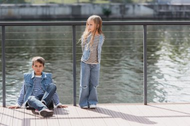 full length of well dressed kids in denim vests and jeans posing next to fence on river embankment clipart