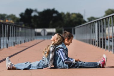 side view of happy preteen kids in denim clothes sitting back to back on river embankment 