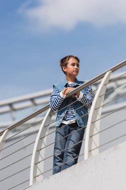 low angle view of preteen boy in trendy denim outfit posing near metallic fence on embankment  clipart