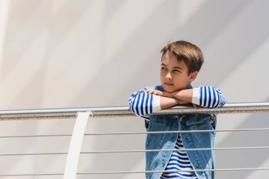 stylish boy in denim vest and long sleeve shirt standing near metallic fence clipart