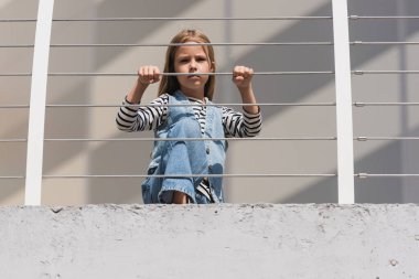 low angle view of stylish kid in denim outfit looking at camera near metallic fence 