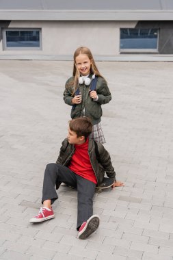 full length of preteen boy in trendy bomber jacket sitting on penny board near cheerful girl 