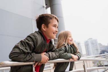 happy preteen kids in bomber jackets leaning on metallic handrails near mall 