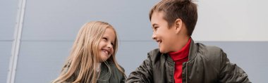 happy preteen kids in bomber jackets looking at each other while standing near mall, banner 