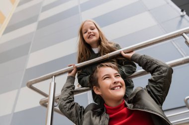 low angle view of cheerful preteen kids in bomber jackets leaning on metallic handrails near mall 