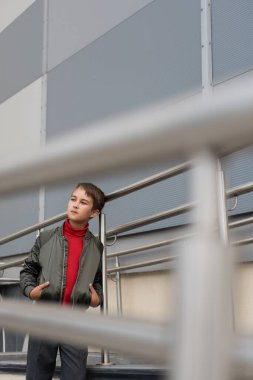 İyi giyimli, havalı bomba ceketli, ön planda metalik tırabzanların yanında elleri ceplerinde poz veren çocuk. 
