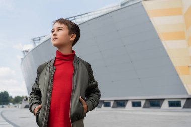 İyi giyimli, havalı bomba ceketli, kırmızı balıkçı yakalı, elleri ceplerinde alışveriş merkezinin yakınında poz veren çocuk. 
