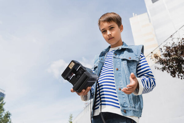 low angle view of well dressed preteen boy in denim clothes holding vintage camera outside 
