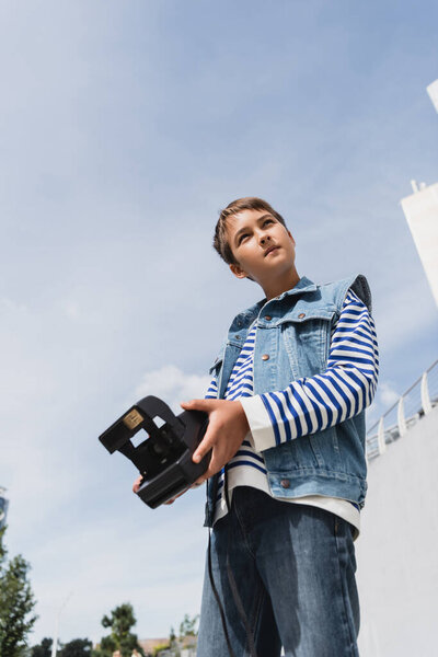 low angle view of well dressed preteen boy in denim outfit holding vintage camera outside 
