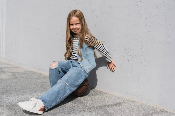 full length of cheerful girl in denim vest and blue jeans sitting on basketball near wall of mall building
