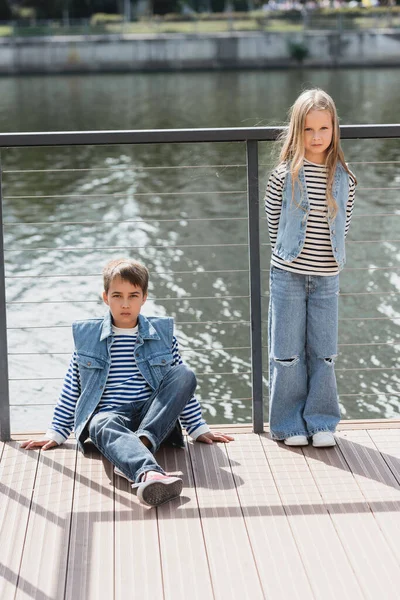 full length of well dressed kids in denim vests and jeans posing next to fence on embankment of river