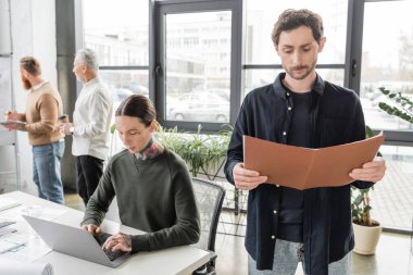 Ofisinde kağıt klasörü olan iş arkadaşının yanındaki bilgisayarı kullanan dövmeli bir işadamı. 