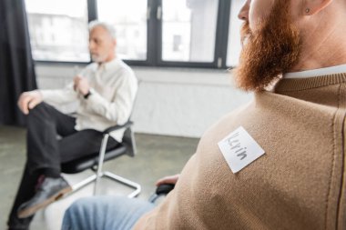 Bearded man with name sticker sitting at group therapy session in rehab center  clipart
