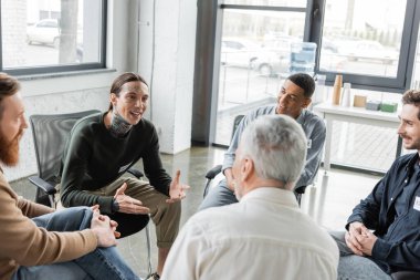 Cheerful tattooed man with alcohol addiction talking to interracial group during therapy in rehab center  clipart