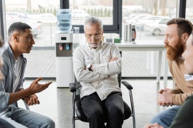 Orta yaşlı üzgün bir adam rehabilitasyon merkezinde alkolikler toplantısında ırklar arası insanların yanında oturuyor. 