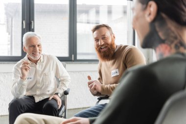 Cheerful people looking at blurred tattooed man during alcoholics meeting in rehab center  clipart