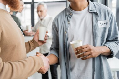 Interracial men with paper cups shaking hands during alcoholics meeting in rehab center  clipart