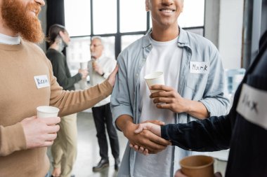 Smiling interracial men with paper cups shaking hands during alcoholics meeting in recovery center  clipart