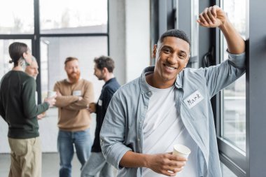 Smiling african american man holding paper cup near blurred group during alcoholics meeting in rehab center  clipart