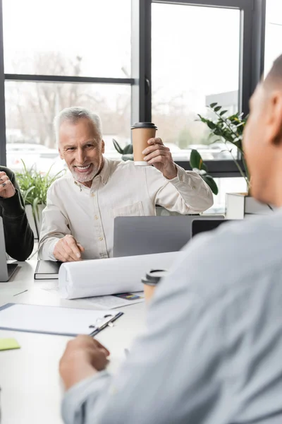 成熟したビジネスマンはオフィスでアフリカ系アメリカ人の同僚をぼやけて近くに行くためにコーヒーを保持 — ストック写真