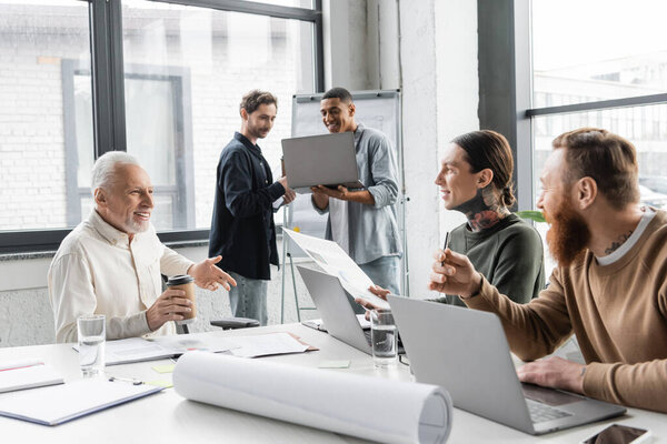 Cheerful businessmen talking near devices and papers during presentation in office 
