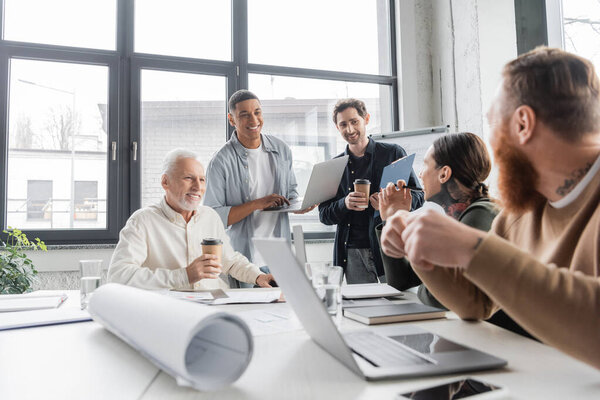 Cheerful multiethnic businessmen with coffee to go using gadgets during training in office 