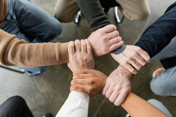 Top view of multiethnic people with alcohol addiction joining hands during group therapy in rehab center 