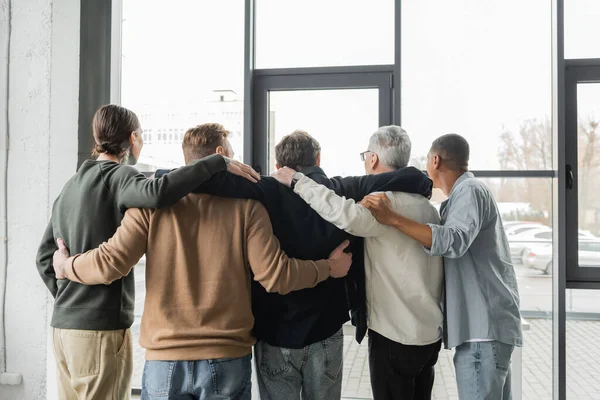 Back view of interracial group of anonymous alcoholics hugging during meeting in rehab center