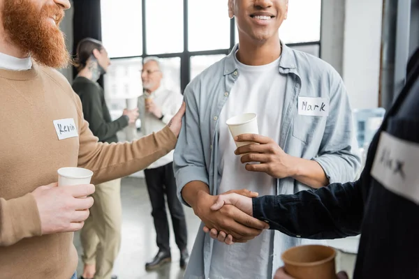 Smiling interracial men with paper cups shaking hands during alcoholics meeting in recovery center