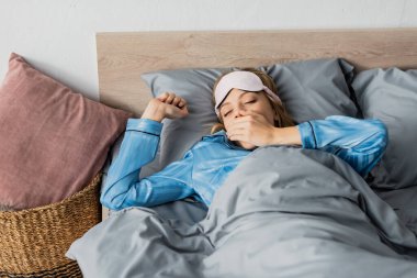 ipek pijama ve gece maskeli uykulu kadın yatakta uzanırken ağzını esnetiyor ve örtüyor. 