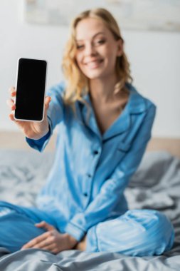 Mavi pijamalı neşeli kadın elinde boş ekranlı akıllı telefon tutuyor. 