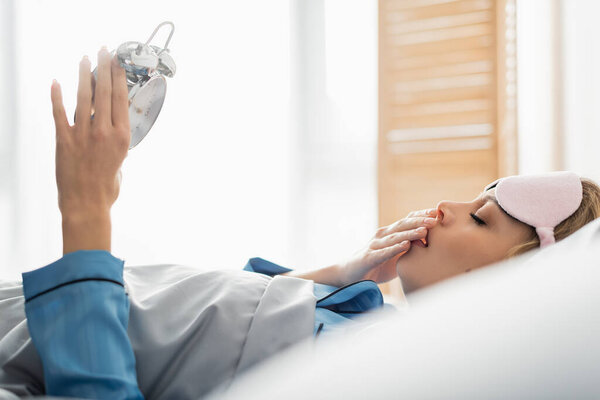 вид сбоку молодой женщины в маске для сна, зевающей, держа в руках будильник по утрам 