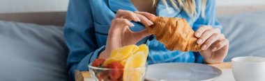 cropped view of young woman in blue pajama holding fresh croissant near tray while having breakfast in bed, banner  clipart