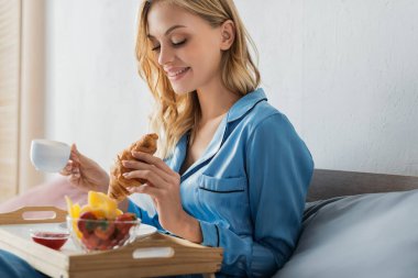 Yatakta kahvaltı yaparken elinde kahve ve kruvasan tutan pijamalı gülümseyen genç kadın. 