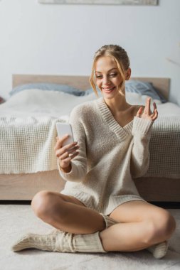 Çağdaş yatak odasındaki akıllı telefondan kazak giyen neşeli genç bir kadın. 