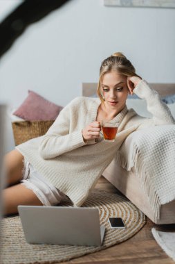 young blonde woman in sweater holding glass cup with tea and looking at laptop in bedroom  clipart