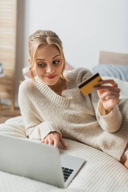Yatak odasında online alışveriş yaparken elinde kredi kartı tutan ve laptop kullanan neşeli bir kadın. 