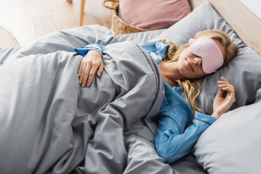 Pembe uyku maskeli ve mavi pijamalı sarışın kadın yatakta dinleniyor. 