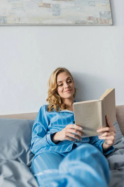 Ευχαριστημένη Νεαρή Γυναίκα Μπλε Μεταξωτό Βιβλίο Ανάγνωσης Νυχτικών Στην Κρεβατοκάμαρα — Φωτογραφία Αρχείου